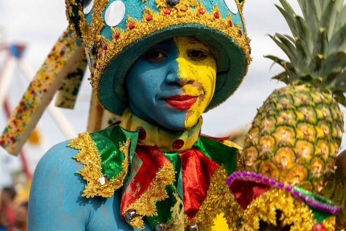 Person dressed in colorful costume holding a pineapple at a festival or carnival, face painted with blue and yellow, wearing a decorated hat.