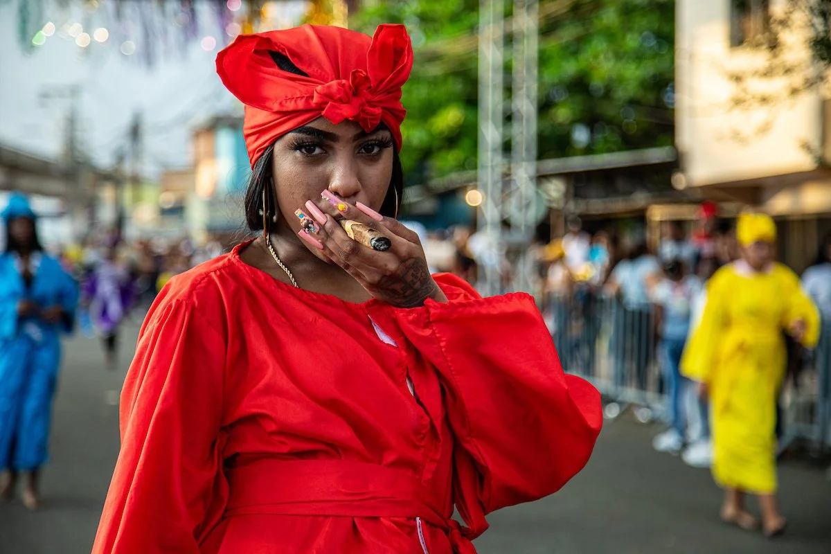 A woman in a red outfit and matching headscarf smoking a cigarette at a street event with many people in the background.