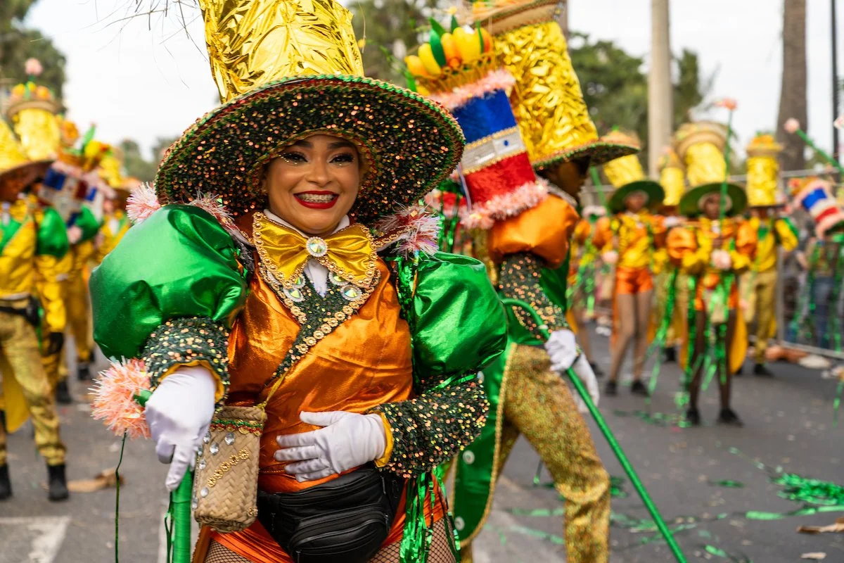 Woman dressed in colorful carnival costume with large hat, green and gold outfit, and white gloves, smiling during parade with other performers in vibrant costumes in background.