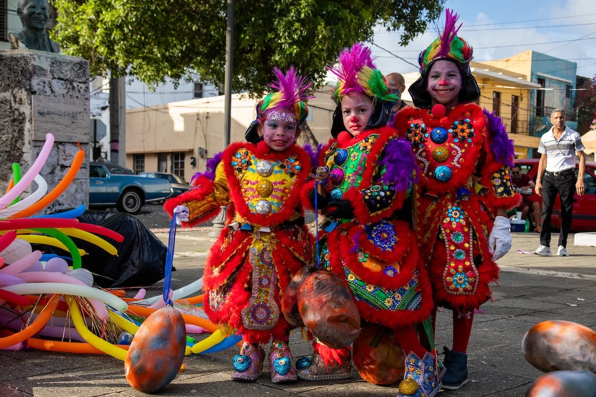 Three children dressed in colorful, elaborate costumes with face paint, participating in a street carnival or celebration, surrounded by balloons and street spectators.