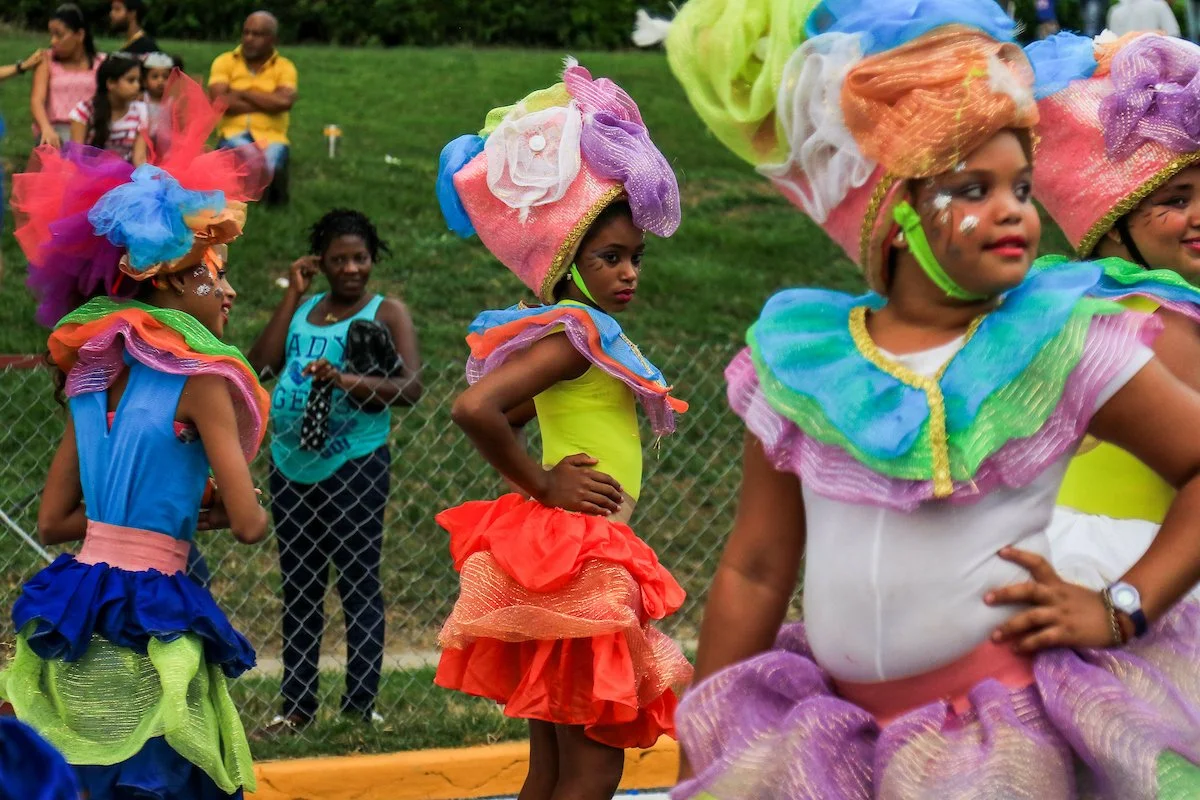 Children dressed in colorful costumes performing in a parade or outdoor event, with some spectators watching in the background.