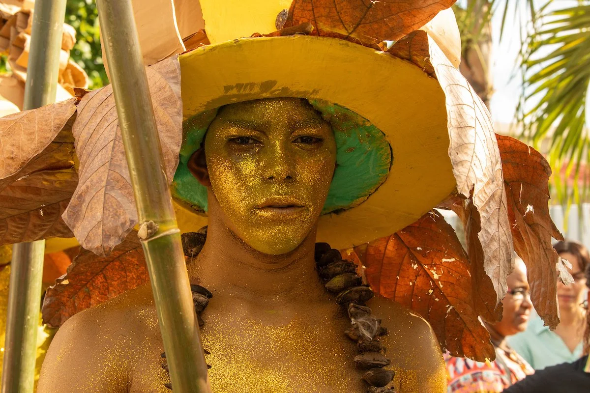 A person dressed as a tropical or fantasy character with glittered gold face paint, wearing a large yellow hat with leaves and a green interior, surrounded by leaves and tropical plants.
