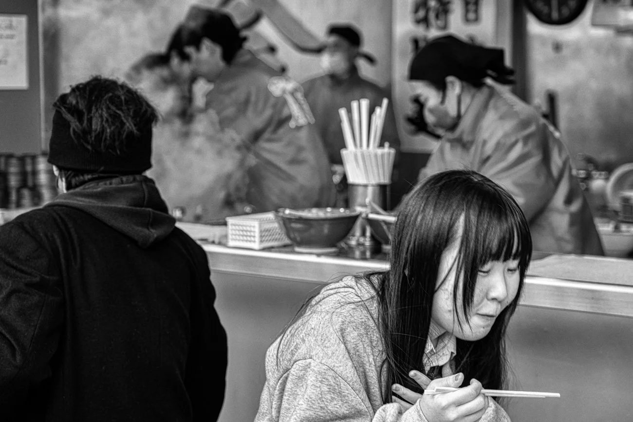 A young woman with long dark hair and a gray sweater using chopsticks in a restaurant with a busy kitchen in the background.