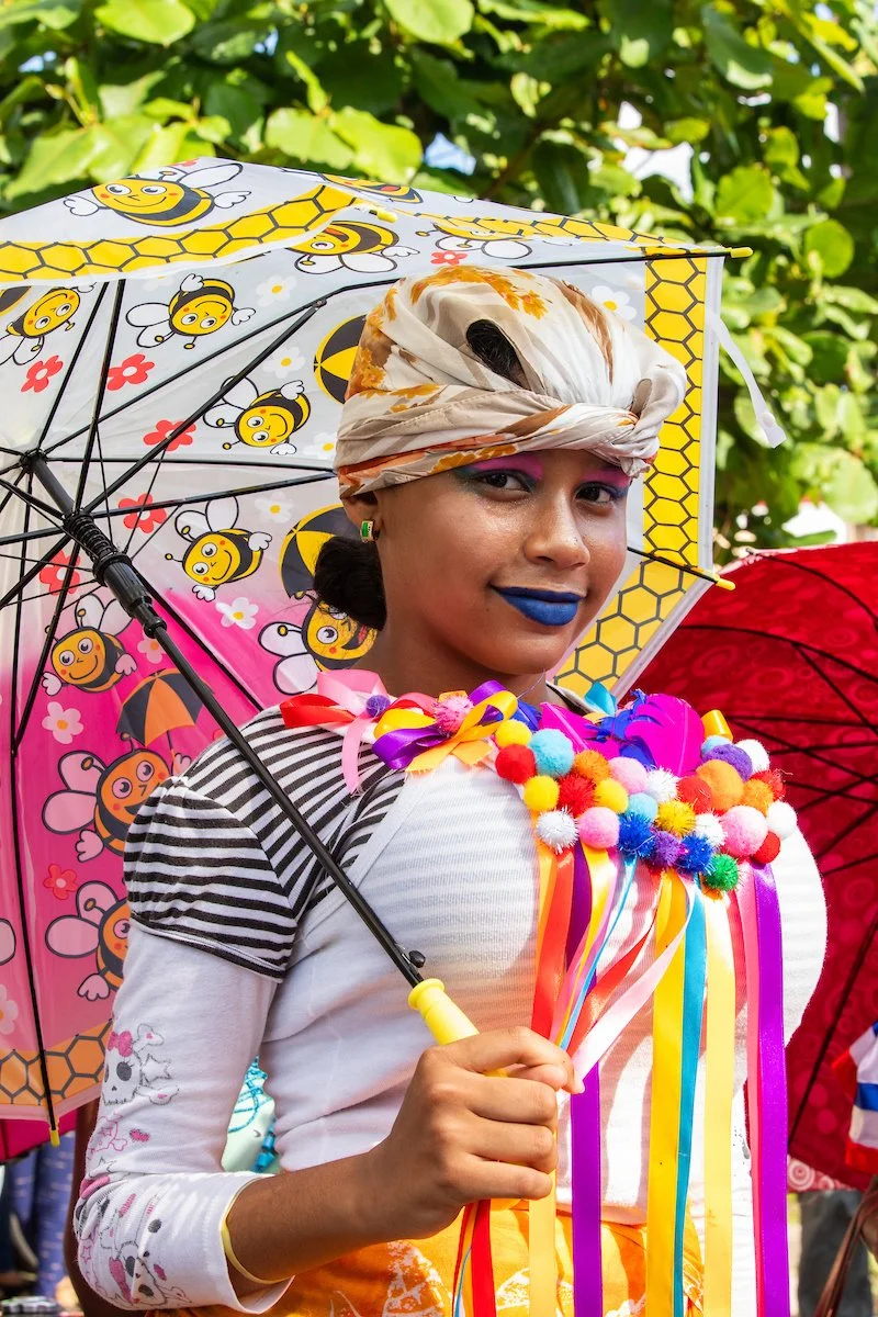 Person in colorful, festive costume holding a decorated umbrella at a parade.