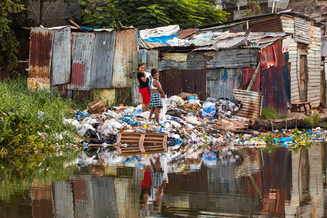 Three children standing on a pile of trash by a waterway, with a makeshift shack made of corrugated metal in the background.