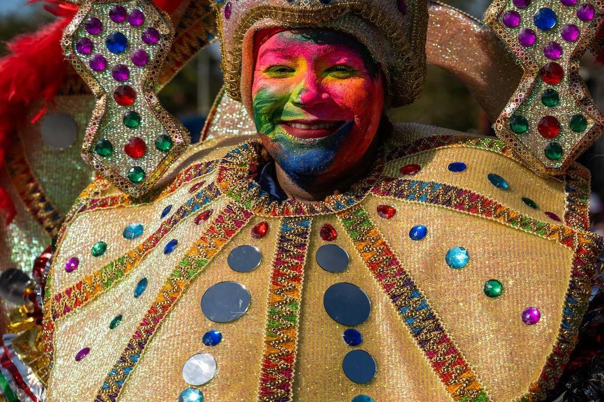 Person smiling with face painted in rainbow colors wearing a colorful, sequined costume with large, shiny, and multi-colored embellishments.