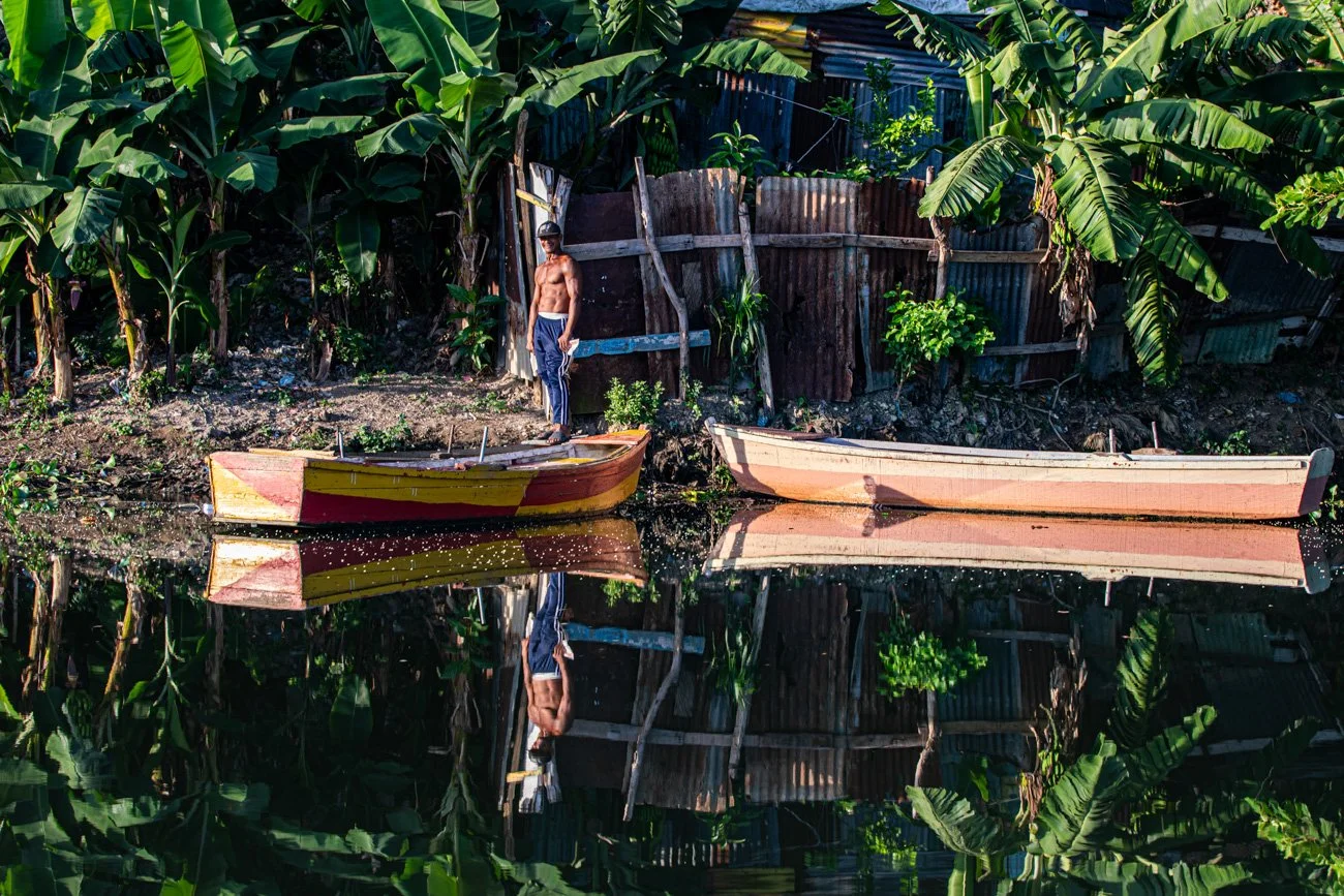 A shirtless man standing on a yellow and red boat by a waterway, with a background of green banana trees and a wooden fence.