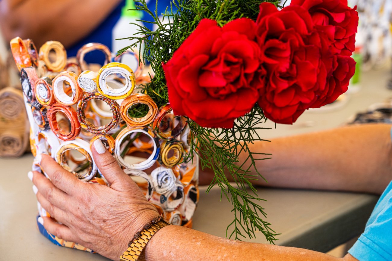 A person arranging a bouquet with three large red carnations and green foliage in a vase covered with rolled paper or pipe cleaner decorations.