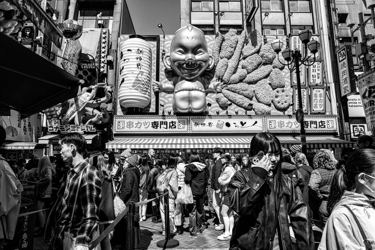 Crowd of people walking in a busy street with large cartoonish statue of a laughing baby on top of a building in the background, decorated with large lanterns and signs in Asian script.