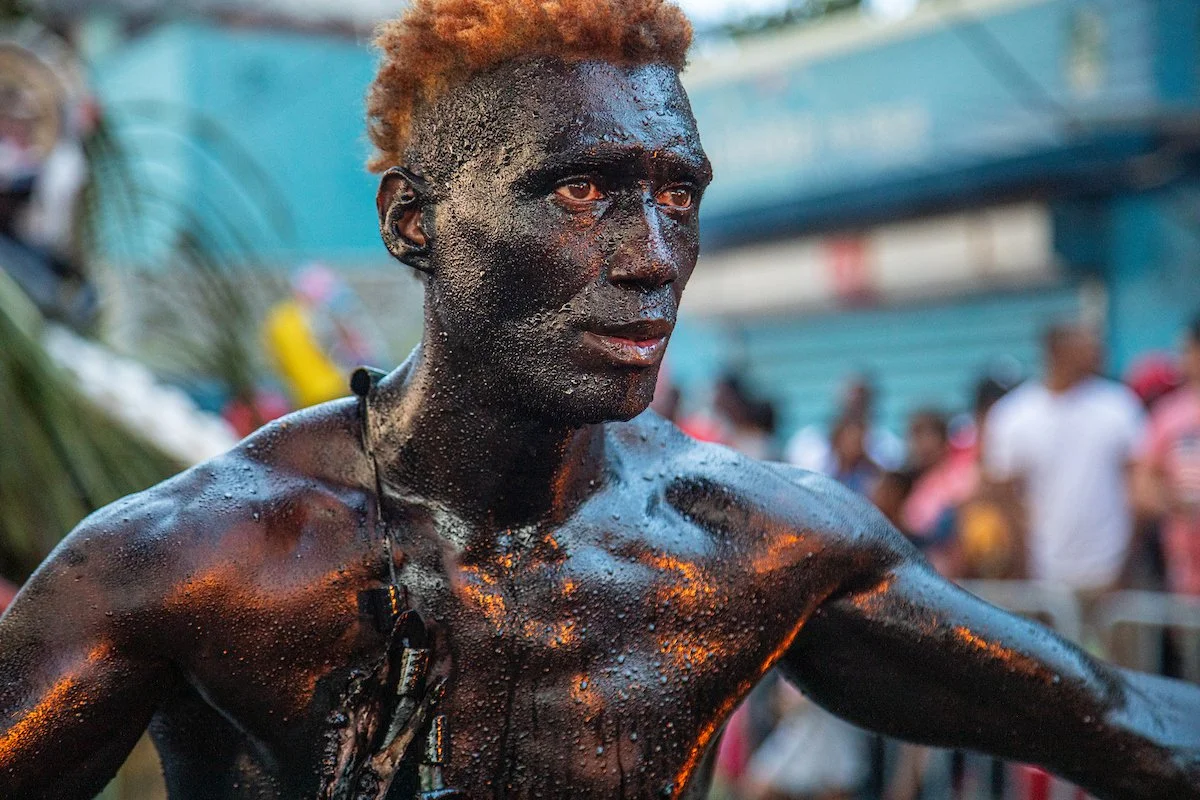 A man covered in a dark, shiny substance, possibly oil or soot, with wet skin and short reddish-brown hair, outdoors with a blurred crowd in the background.