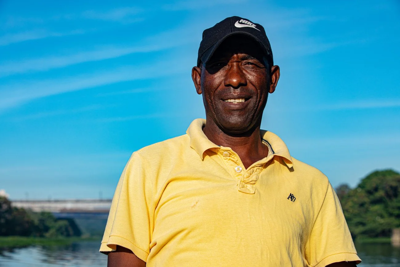 A smiling man wearing a yellow polo shirt and a black Nike cap, standing outdoors near a body of water with a bridge and green trees in the background on a clear, sunny day.