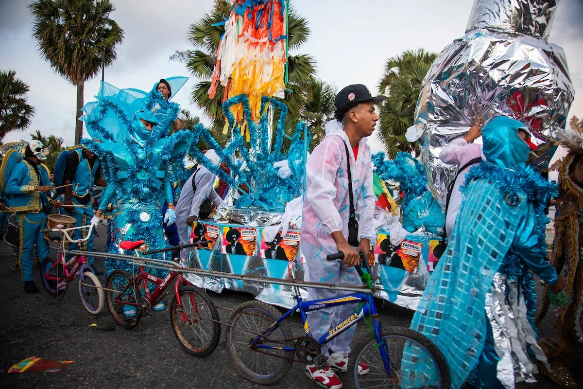 People participating in a parade wearing colorful costumes, some riding decorated bikes and others pushing floats, with palm trees in the background.