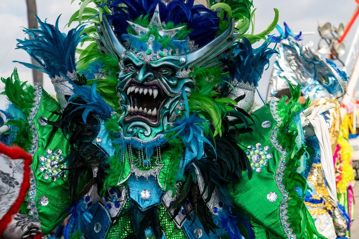 Colorful costume with a fierce, detailed face mask of a demon or mythological creature, surrounded by green, blue, and black feathers and decorative rhinestones, likely part of a parade or cultural festival.