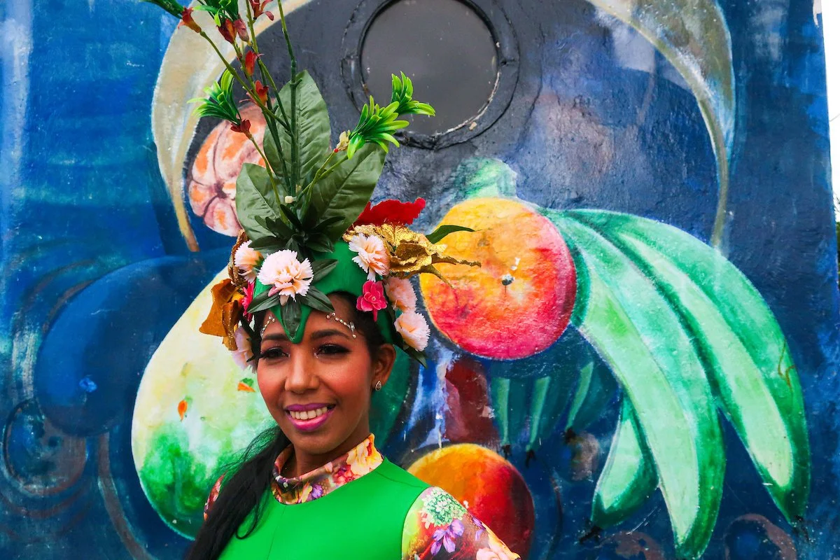 A woman is wearing a vibrant, floral headdress with various flowers and greenery, and a bright green outfit. She is standing in front of a colorful mural featuring stylized fruit, including apples, peaches, and leaves.