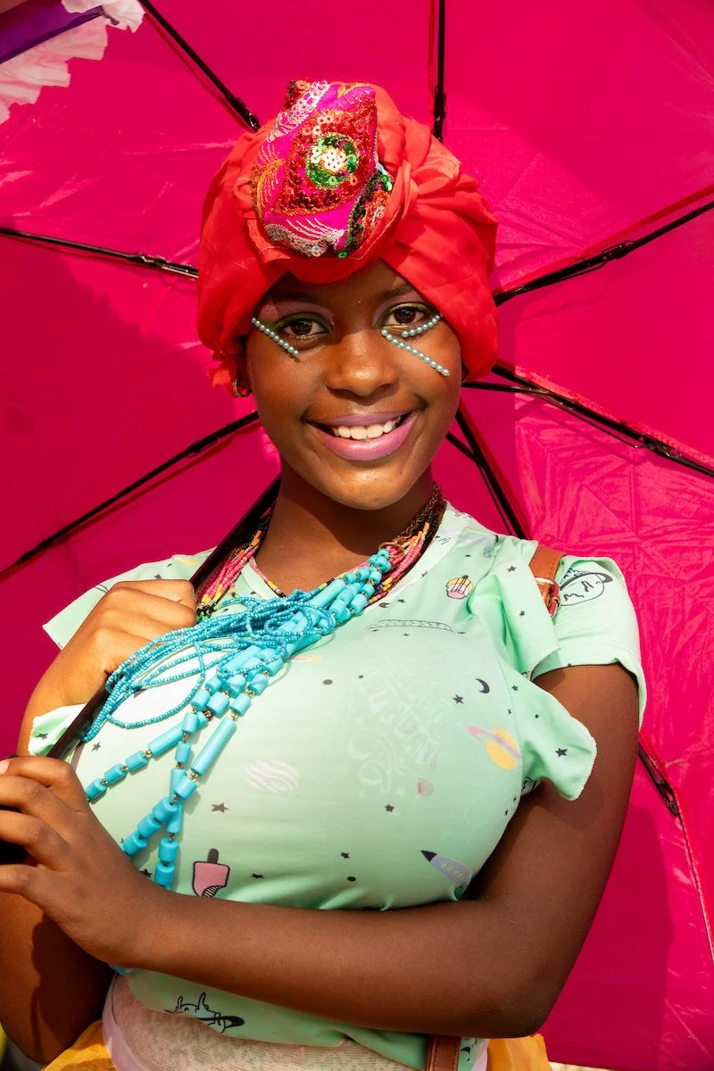 A young woman smiling, holding a pink umbrella. She wears a colorful headwrap with a decorative patch, multiple blue bead necklaces, and face adornments made of small beads under her eyes. She is dressed in a light green shirt with space-themed print