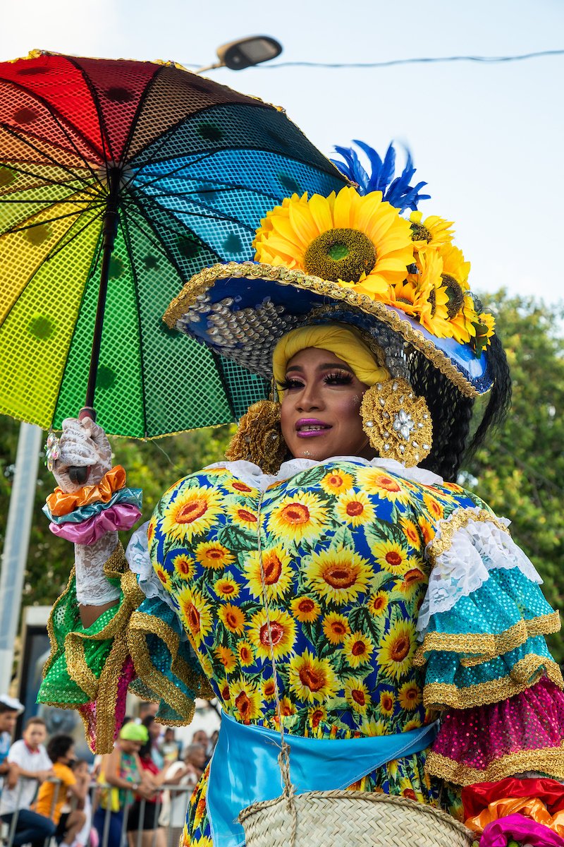 A person dressed in a colorful sunflower-themed costume holding a rainbow umbrella and wearing a large hat decorated with sunflowers and other flowers at a parade or festival.