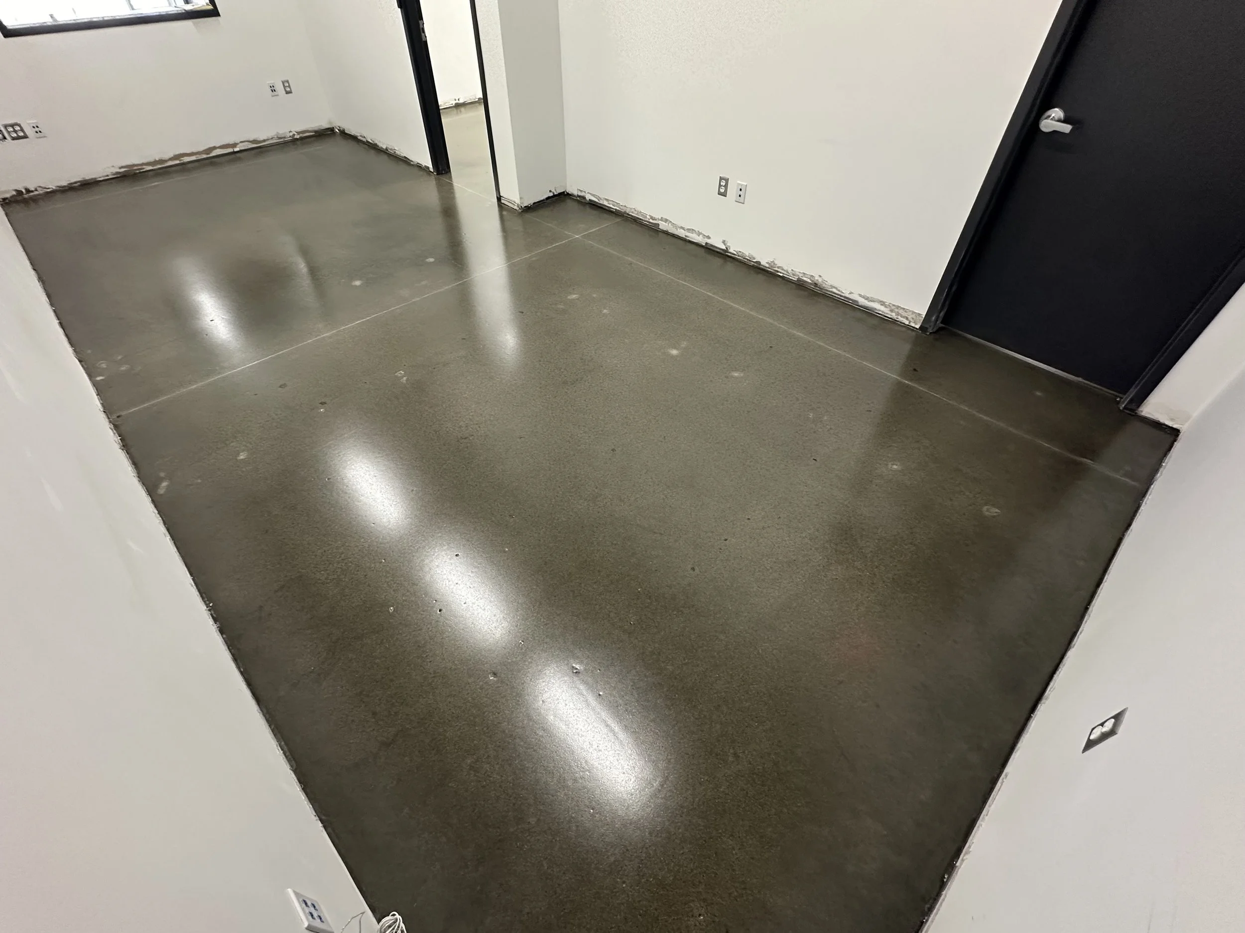 Empty room with polished concrete floors, white walls, and electrical outlets along the walls. An open doorway reveals an adjacent room.