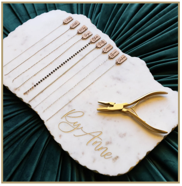 Jewelry display with various necklaces featuring name tags, gold pliers, and a gold cursive sign reading 'Buy Anne' on a white marble tray, surrounded by green fabric