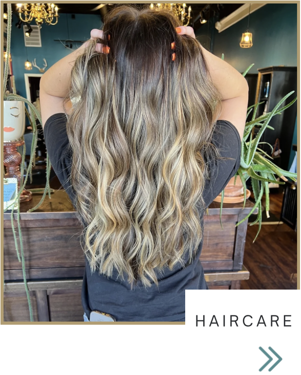 Back view of a woman with long, wavy, blonde-highlighted hair, holding her head with both hands inside a salon with plants and chandeliers.
