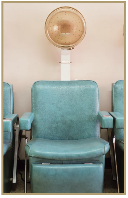 Empty teal salon chairs in front of a hair drying hood in a salon or barbershop.