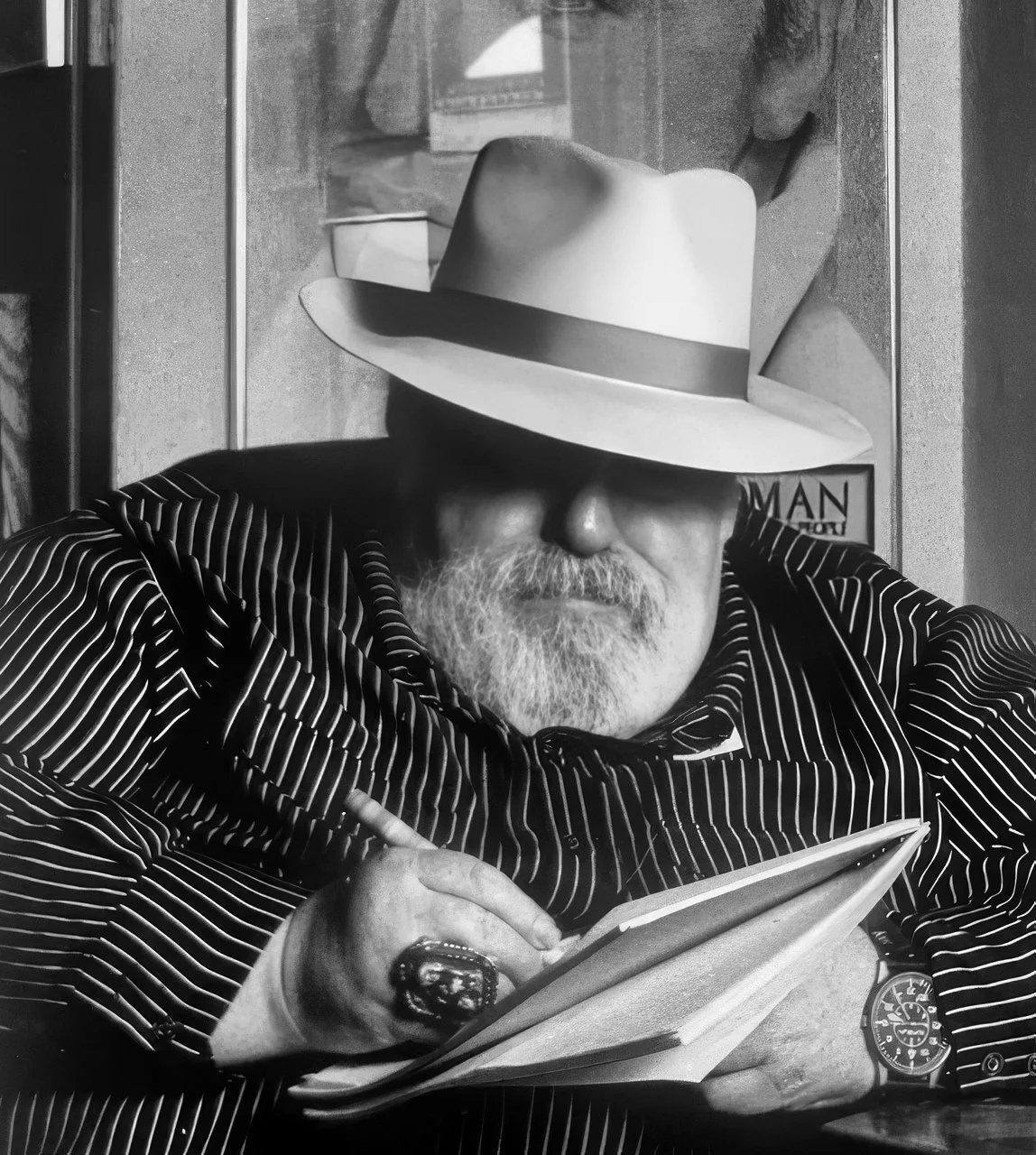 Doc Pomus wearing a wide-brimmed hat, sunglasses, and a pinstripe shirt, is holding a notebook or planner and a pen.