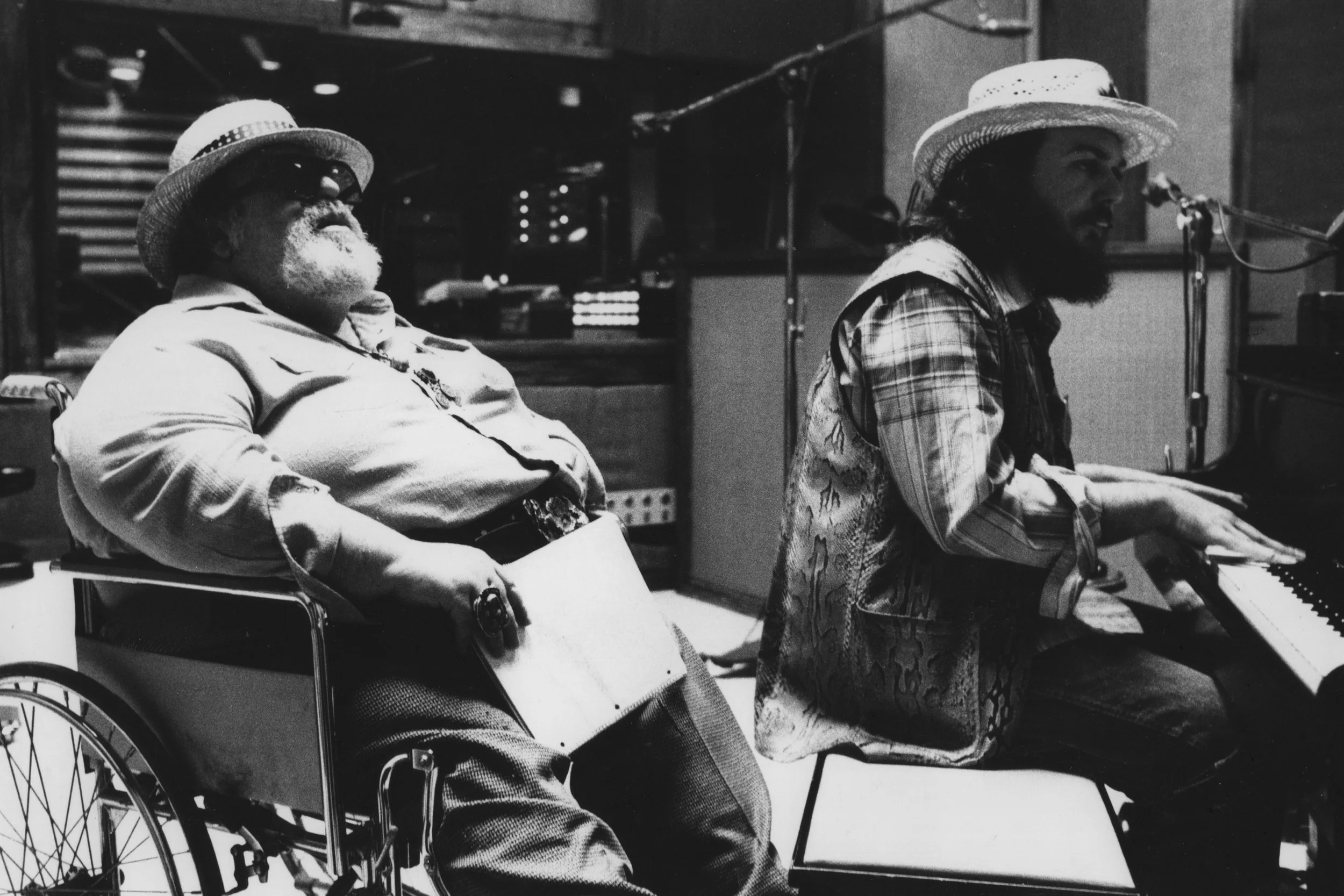 This image captures legendary blues singer and songwriter Doc Pomus in a wheelchair in a recording studio, during his collaboration with Dr. John at the piano in the late 1970s.