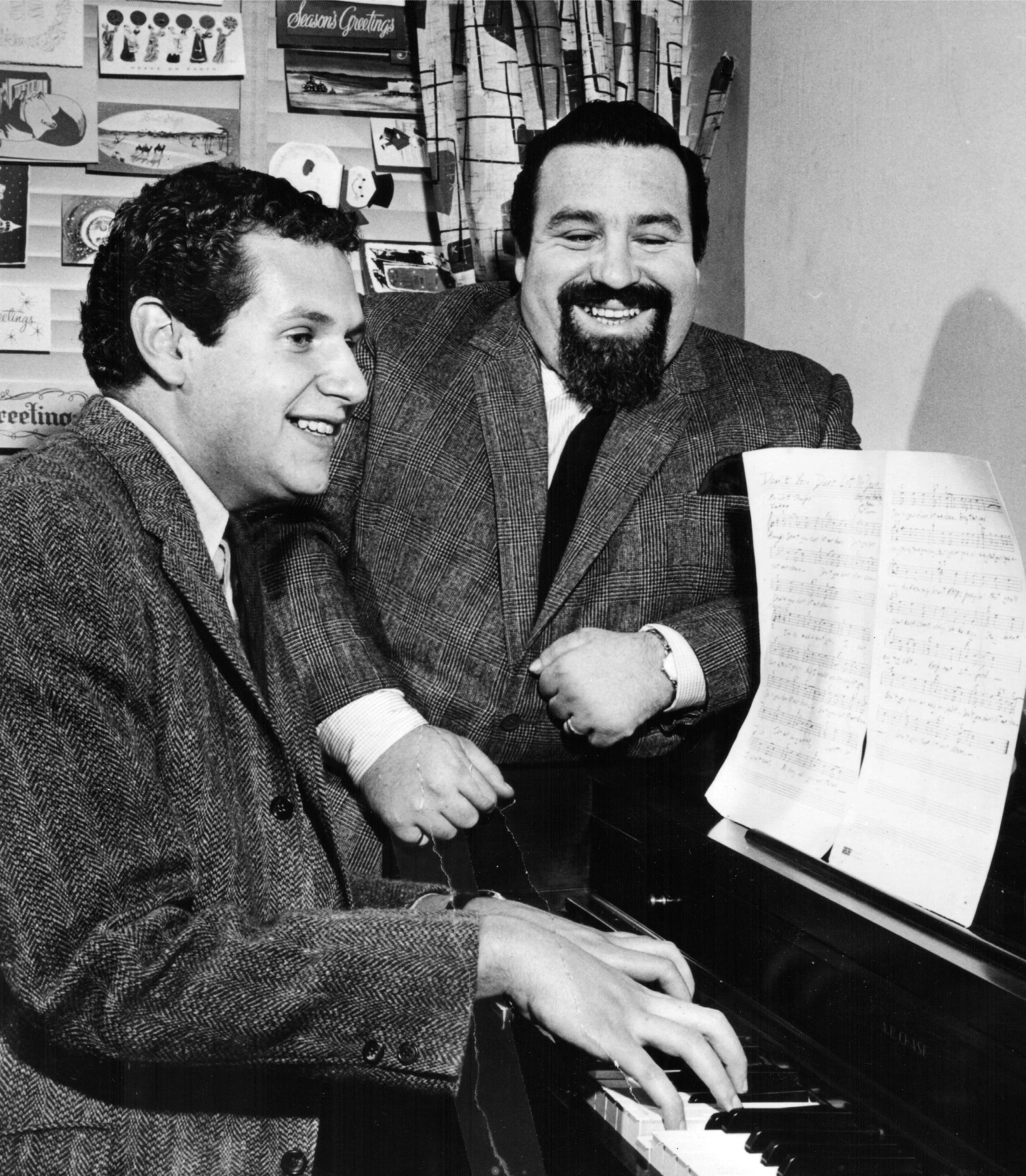 Doc Pomus and Mort Shuman smiling and looking at a sheet of music on a piano in a room decorated with holiday and greeting cards on the wall.