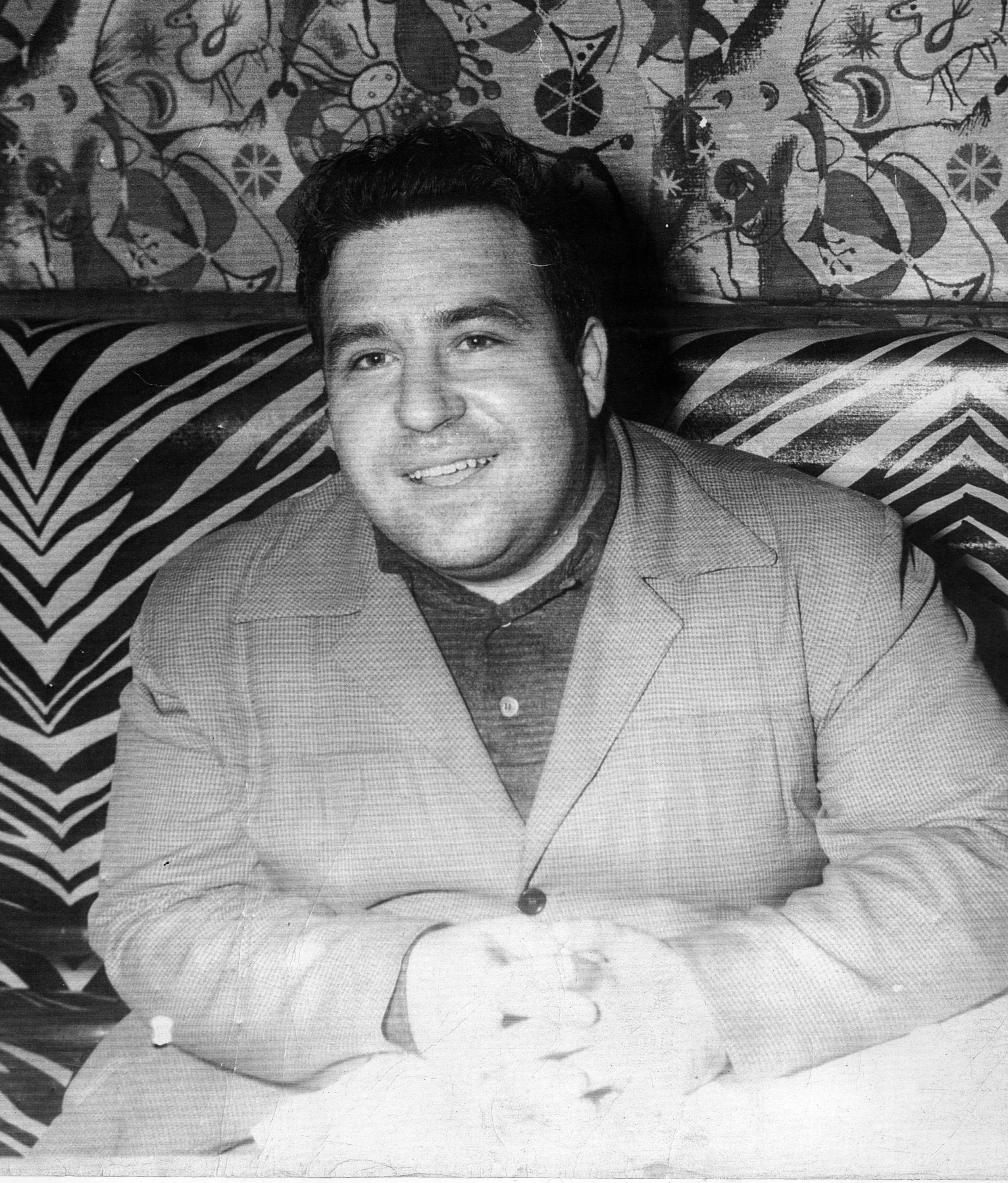Doc Pomus in a suit and dark shirt sitting at a table with patterned decorations in the background.