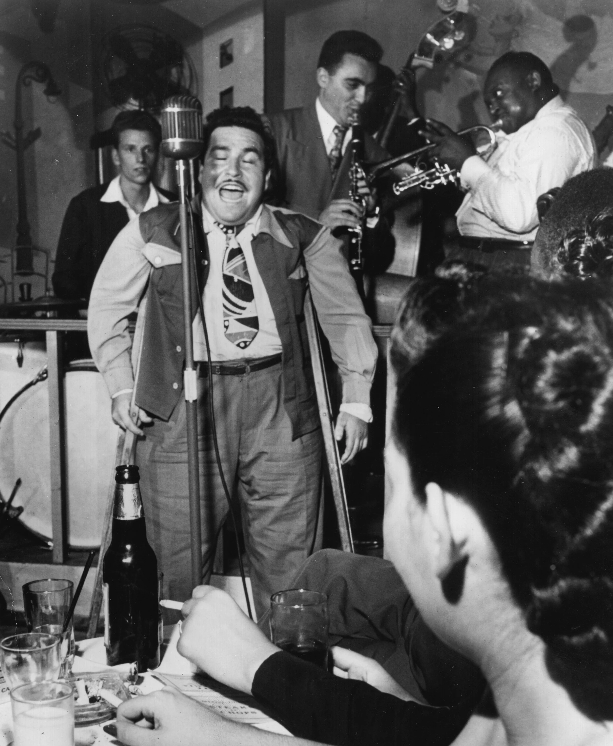 Black and white photo of a lively musical performance in a small venue, with a young Doc Pomus passionately singing at a microphone, surrounded by musicians playing instruments, and an audience member in the foreground watching the performance.