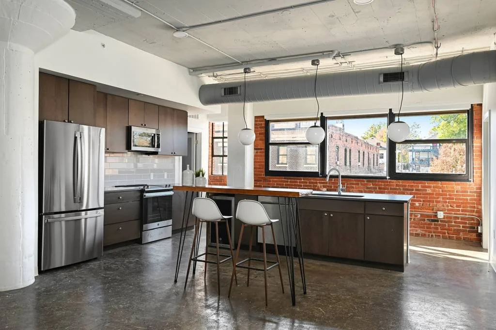 Modern kitchen with stainless steel refrigerator, microwave, and oven, dark brown cabinets, white subway tile backsplash, brick wall accents, large window, black countertop island with two white chairs, pendant lights hanging from an exposed ceiling 