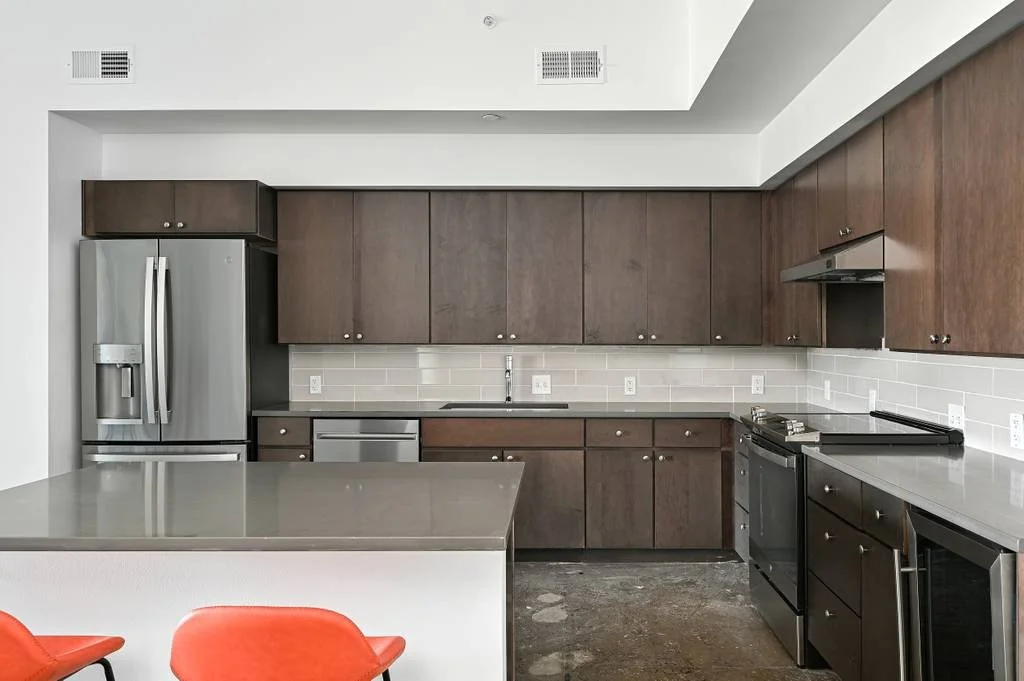 Modern kitchen with brown cabinets, stainless steel refrigerator, stove, and dishwasher. Gray countertops, white subway tile backsplash, and two orange chairs at a gray island.