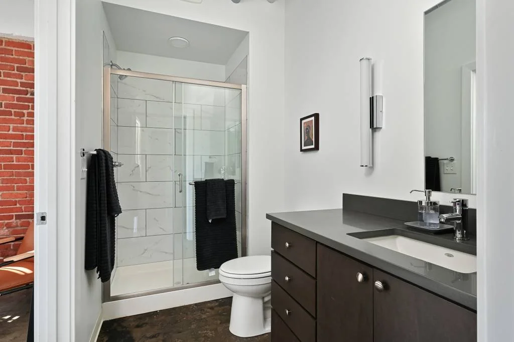Bathroom with a glass-enclosed shower, a white toilet, and a dark wood vanity with a gray countertop and a small sink. Black towels hang on a towel bar and the shower door. There's a framed picture on the wall and a modern vertical light fixture abov