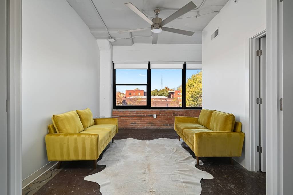 A bright room with white walls, a ceiling fan, large window with a view of trees and buildings outside, two yellow sofas facing each other, a white rug on the dark wooden floor.