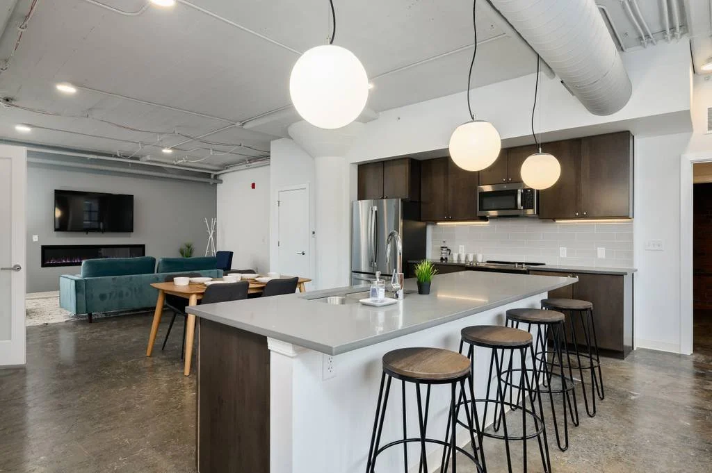 Open kitchen with white countertop island, three bar stools, dark wood cabinets, stainless steel refrigerator, oven, microwave, and gray subway tile backsplash. In the background, a dining table with black chairs, and a living area with a teal sofa a