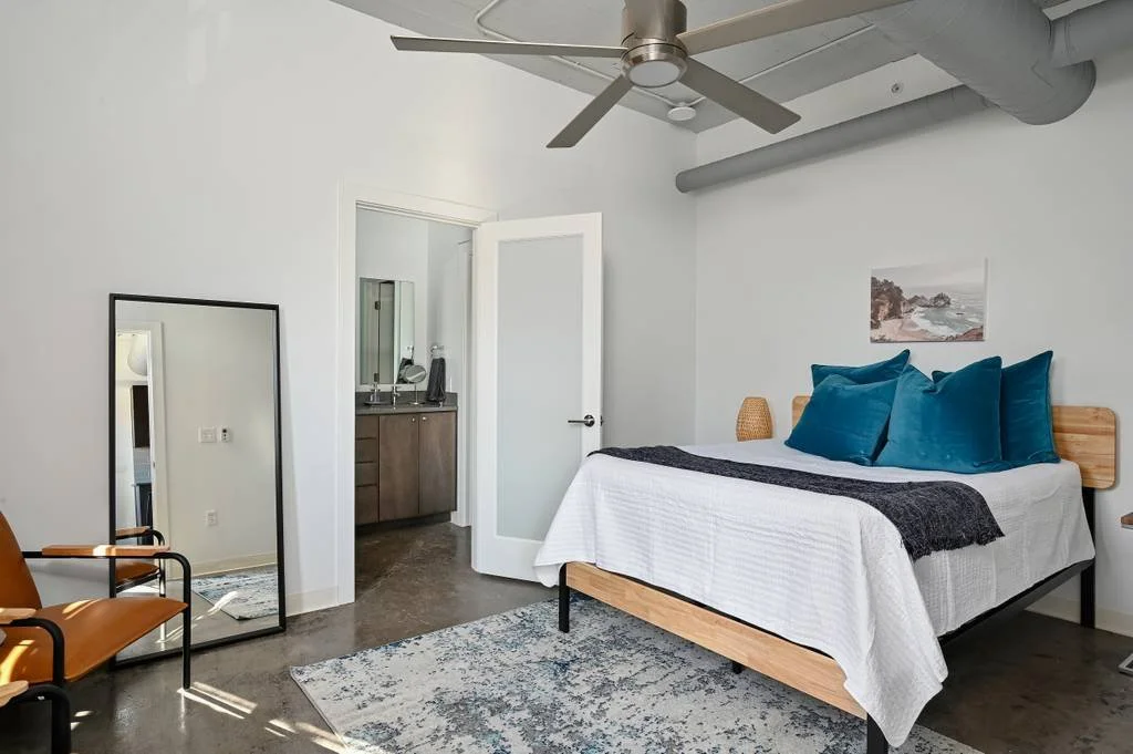 Modern bedroom with a wooden bed, white bedding, and teal pillows. There is a wall art above the bed, a mirror reflecting a chair, and a small woven lamp beside the bed. A ceiling fan is on the ceiling.