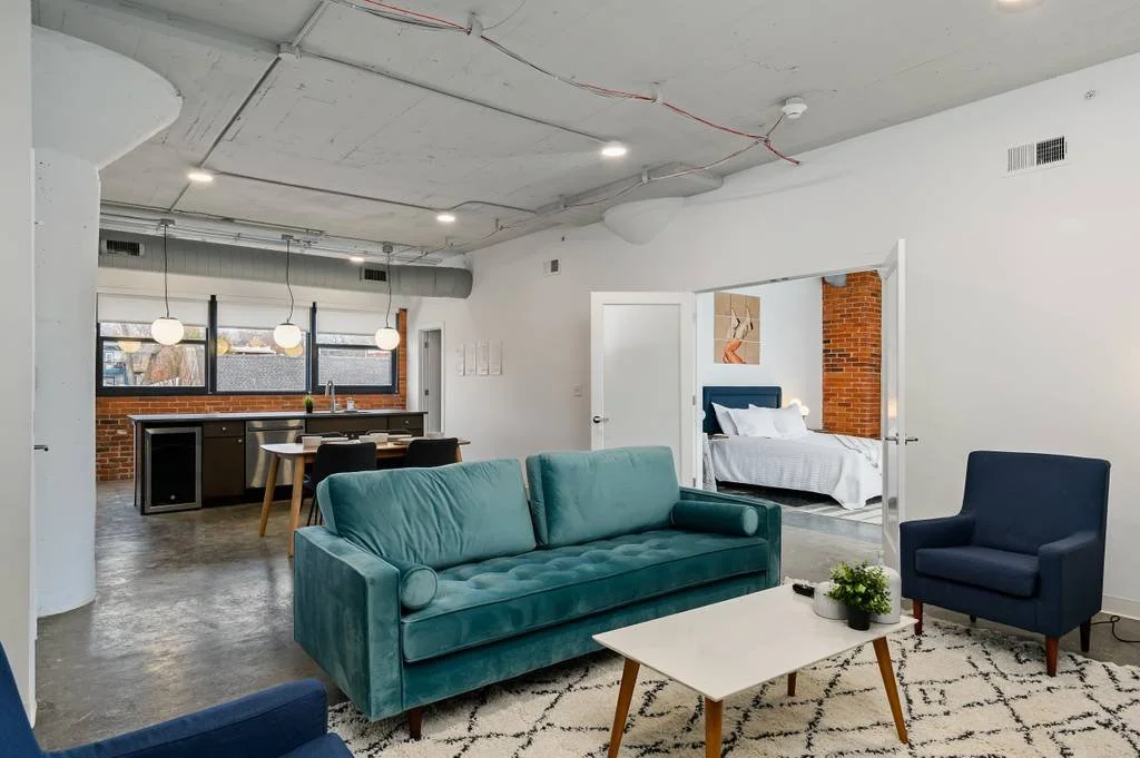 Open-concept living space with a teal velvet sofa, a dark armchair, a white coffee table with plants, and a view into a bedroom with iron bed frame and brick wall, in a modern apartment.