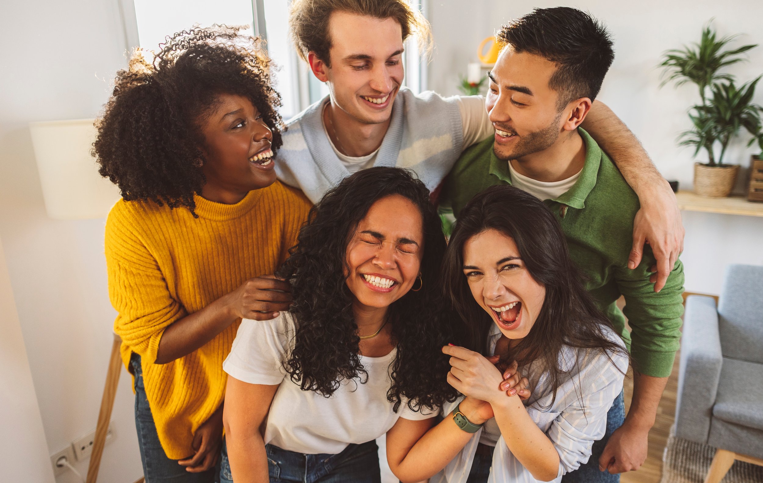 Group of five diverse friends laughing and hugging indoors.