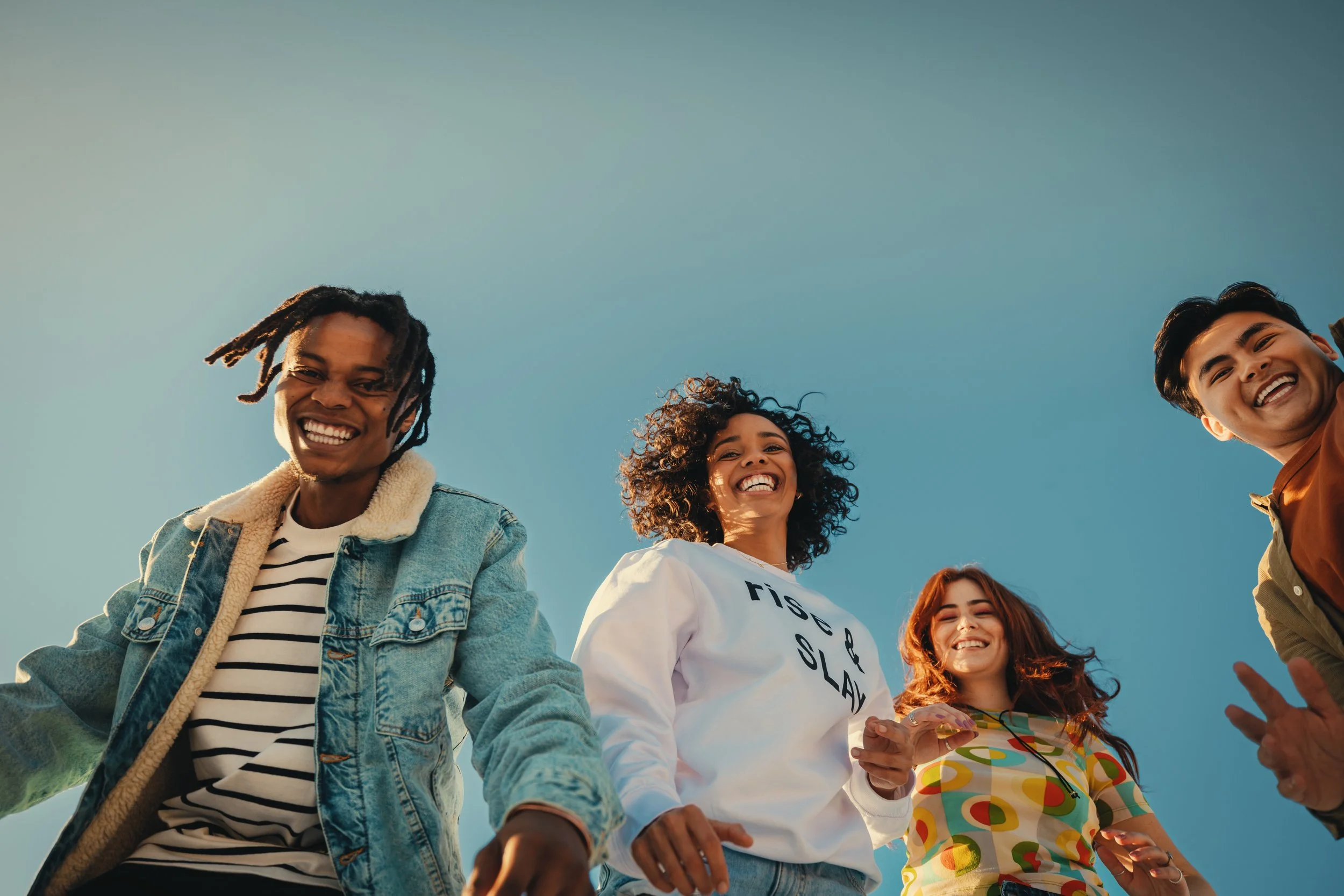 Group of five young adults smiling and laughing outdoors under a clear blue sky.
