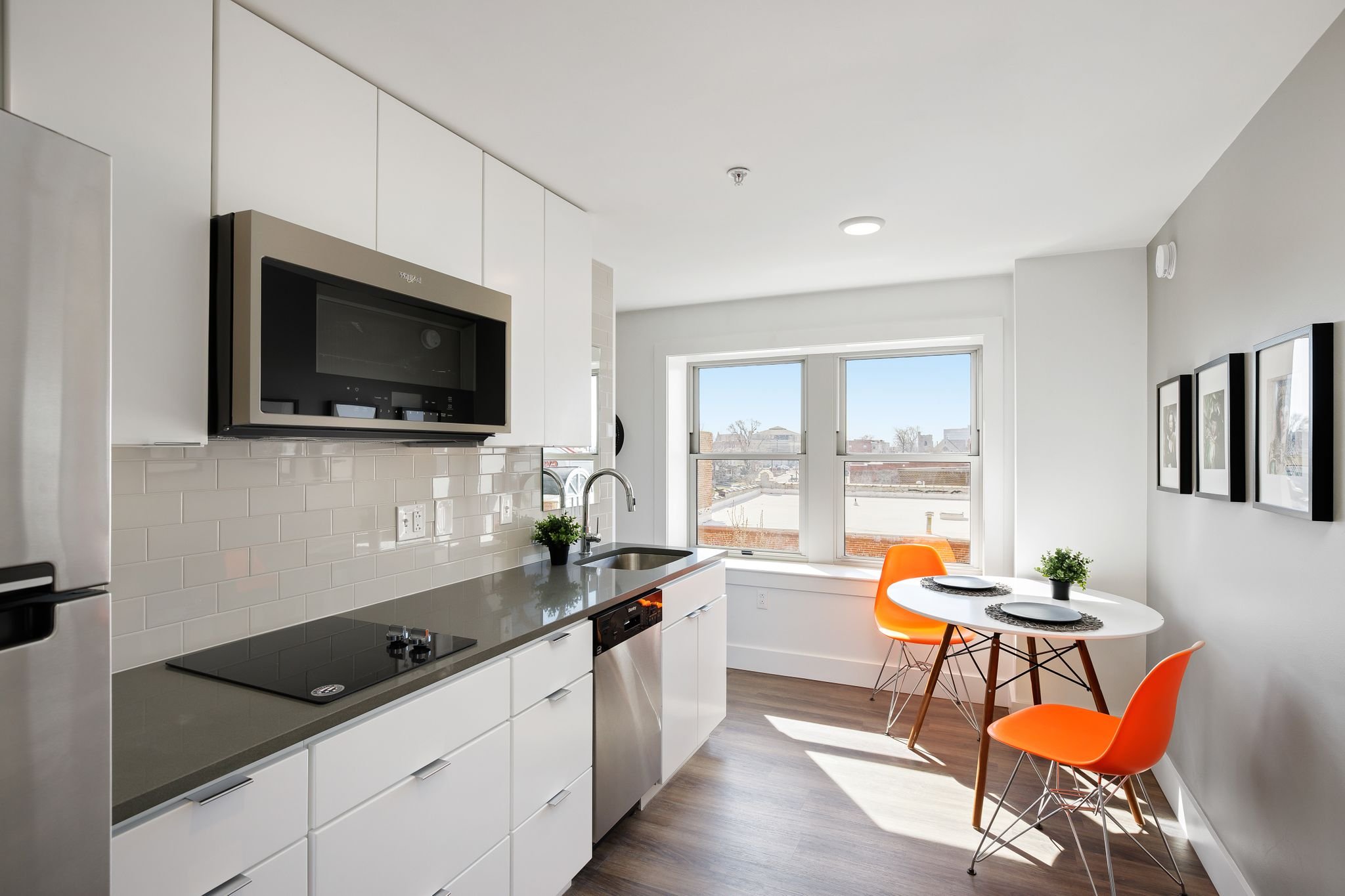 Modern kitchen with white cabinets, gray countertop, stainless steel appliances, a window seat with bright orange chairs, and framed pictures on the wall.