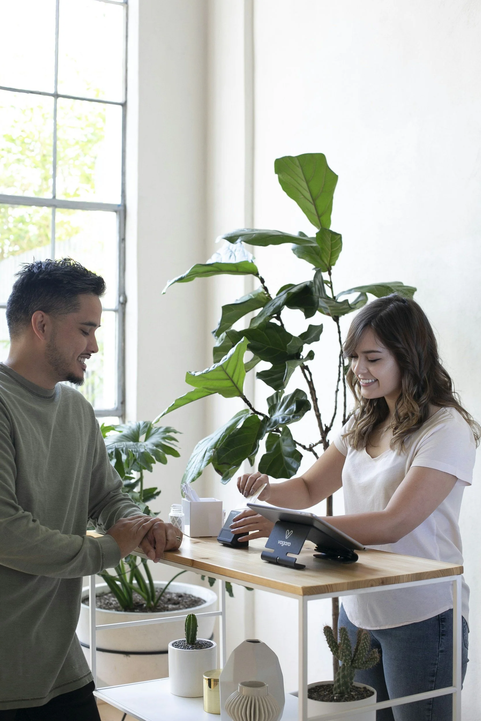 A customer is paying at a retail counter while a smiling cashier processes the transaction. The scene takes place indoors with a large window and green houseplants in the background.