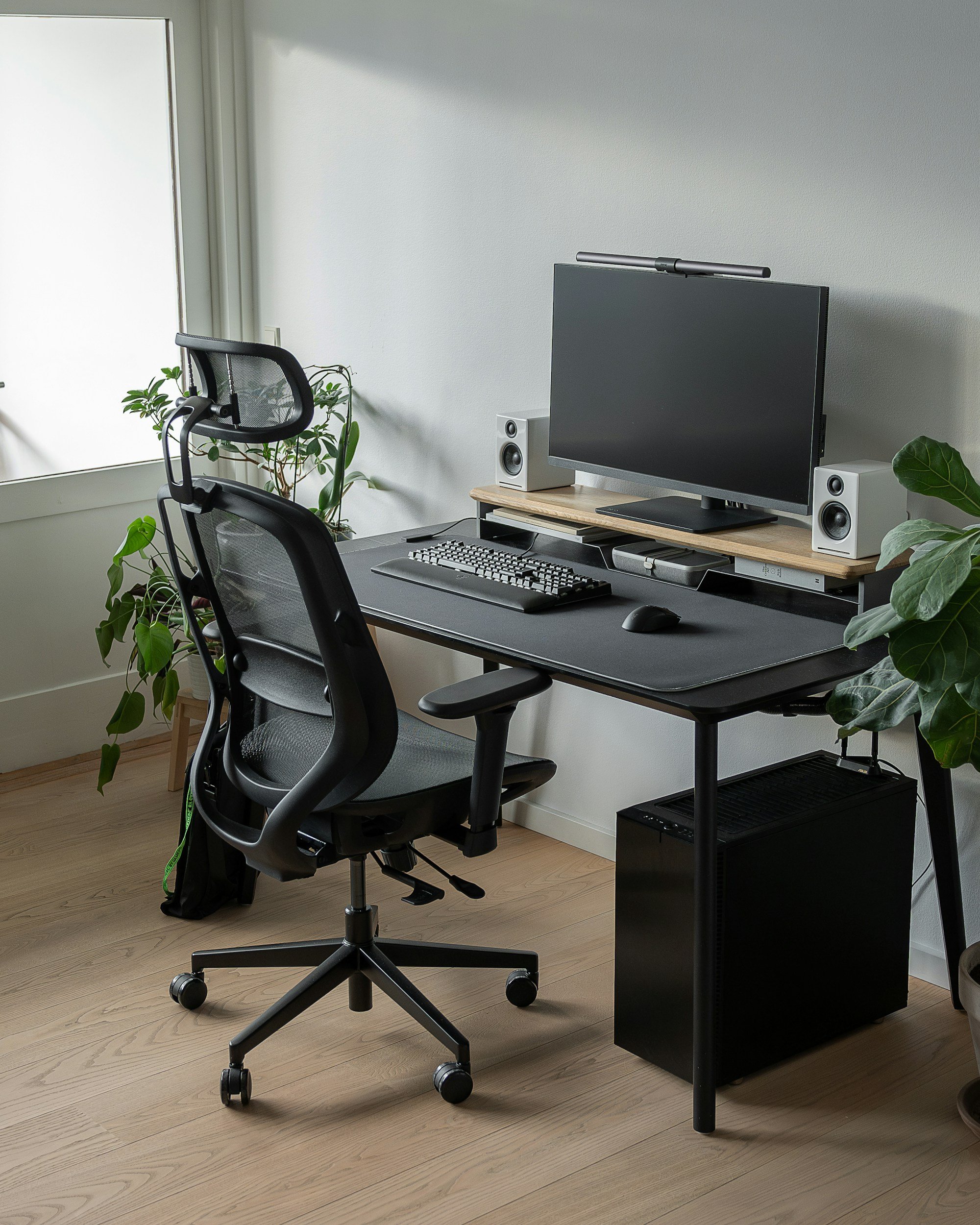 Home office setup with a black desk, dual monitors, a keyboard, mouse, speakers, an ergonomic office chair, and indoor plants near a window.