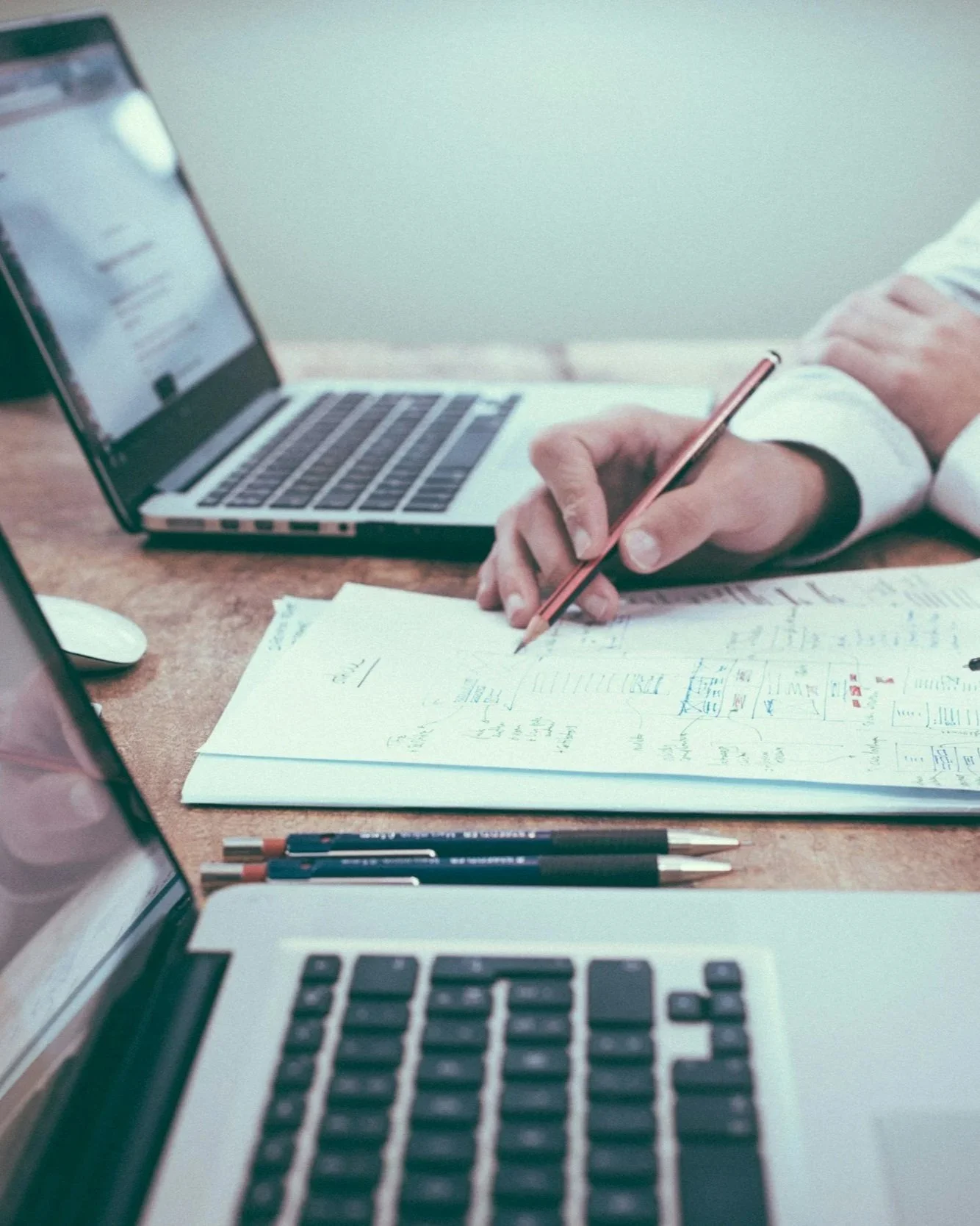 Close-up of a person's hand holding a pencil over handwritten notes and diagrams on paper, with laptops, pens, and a computer mouse on a wooden desk.