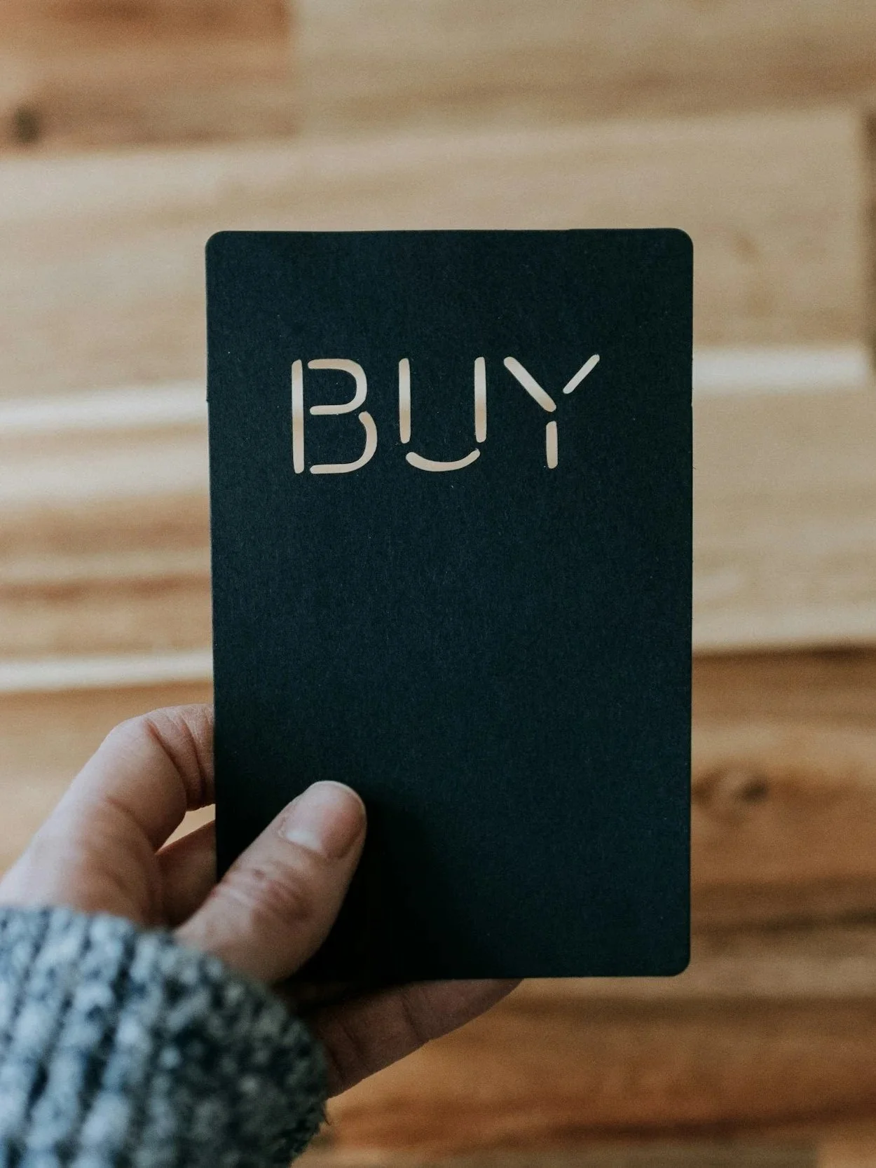 A hand holding a black card with the word 'BUY' cut out in the center.