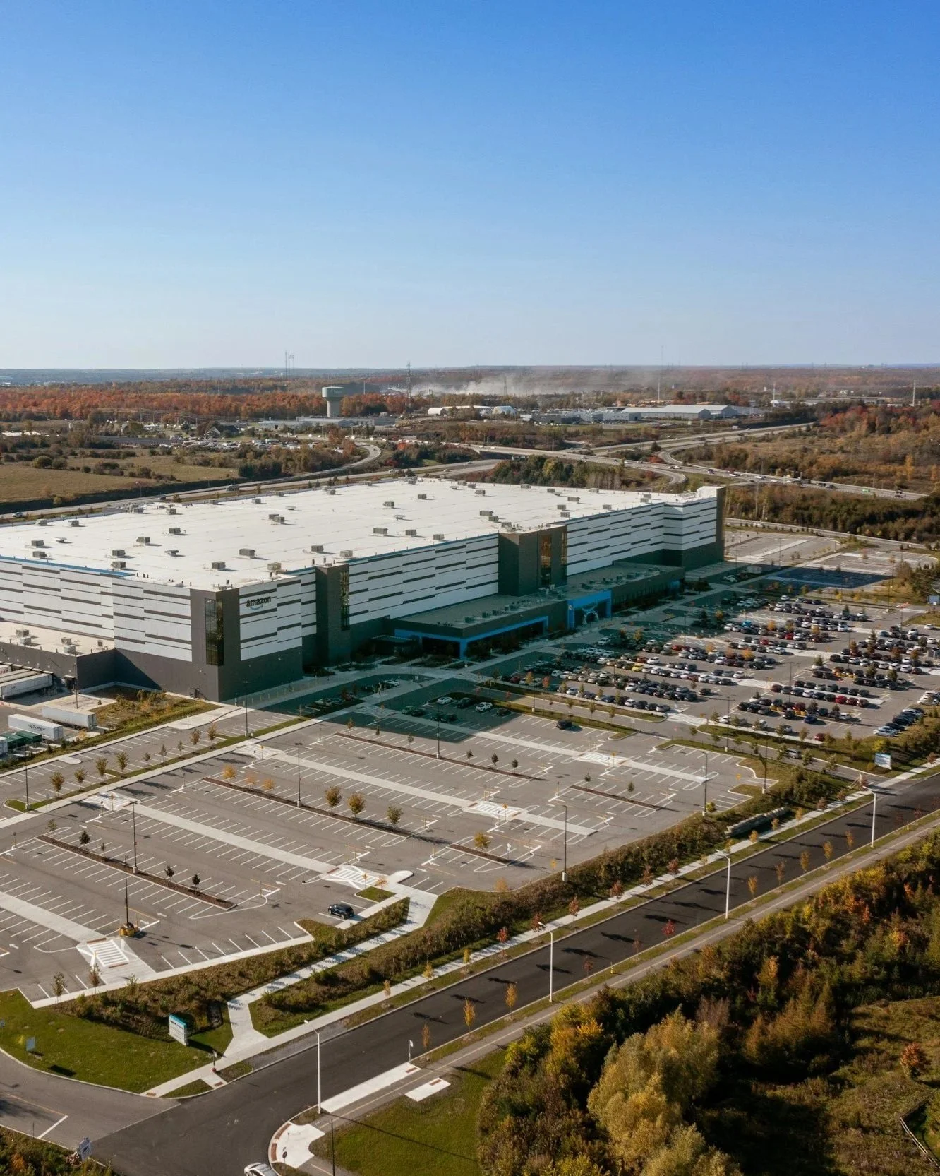 Aerial view of an Amazon distribution center with a large parking lot and surrounding roads, under a clear blue sky.