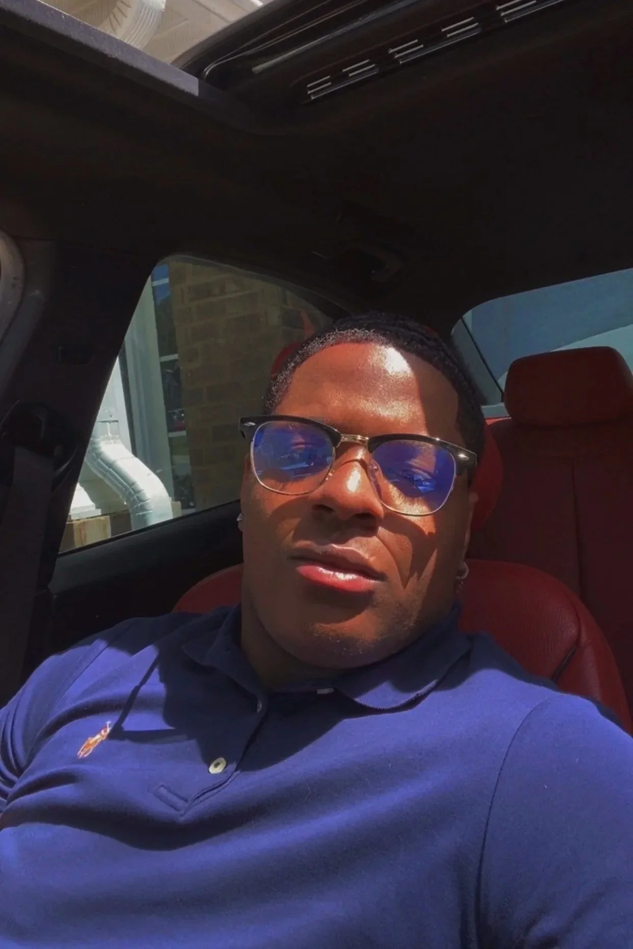 A man wearing sunglasses and a navy blue polo shirt sitting in the back seat of a car, with sunlight shining on his face.