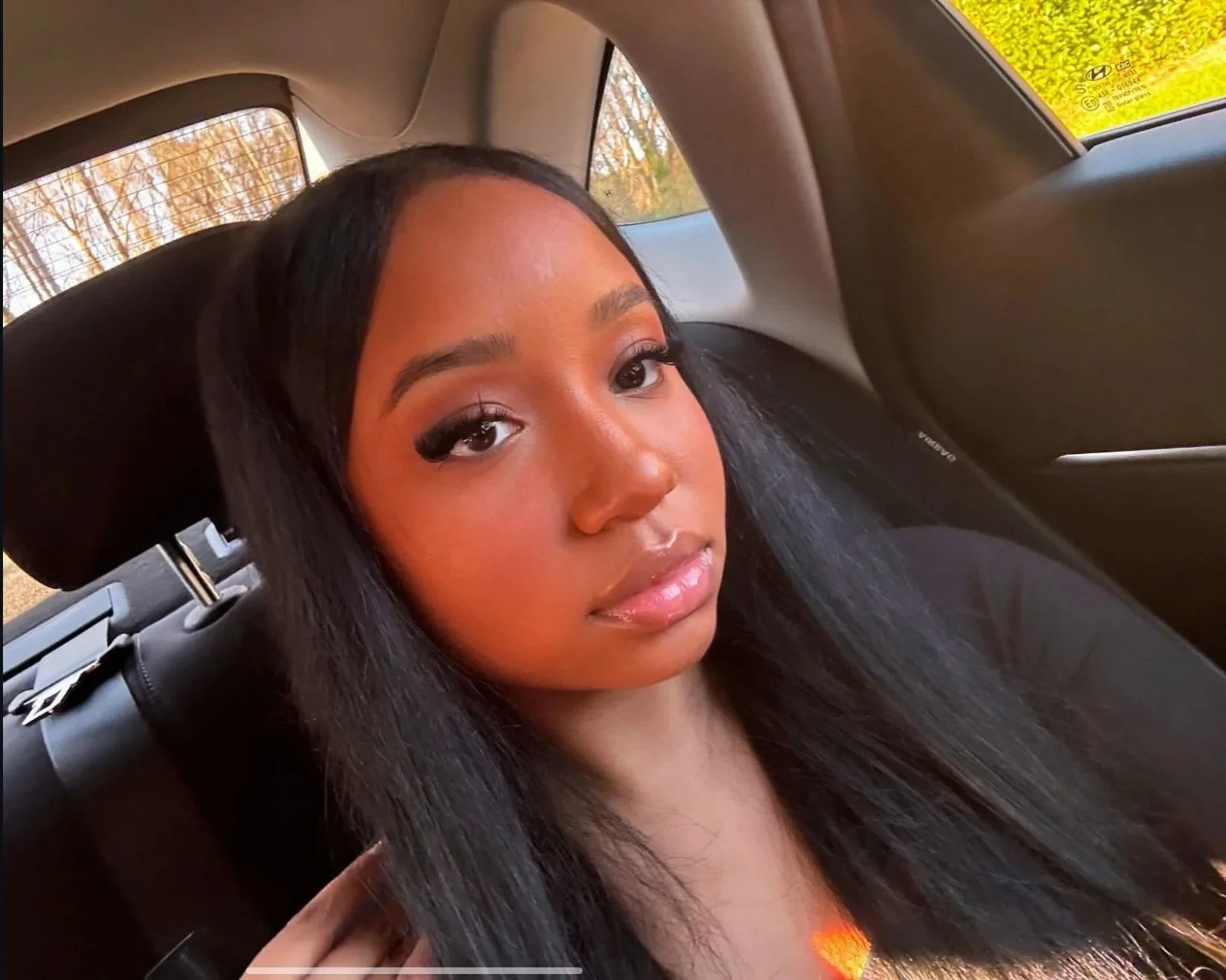A young woman with long, straight black hair, makeup, and glossy lips, sitting in the backseat of a car with a window showing trees and sunlight outside.