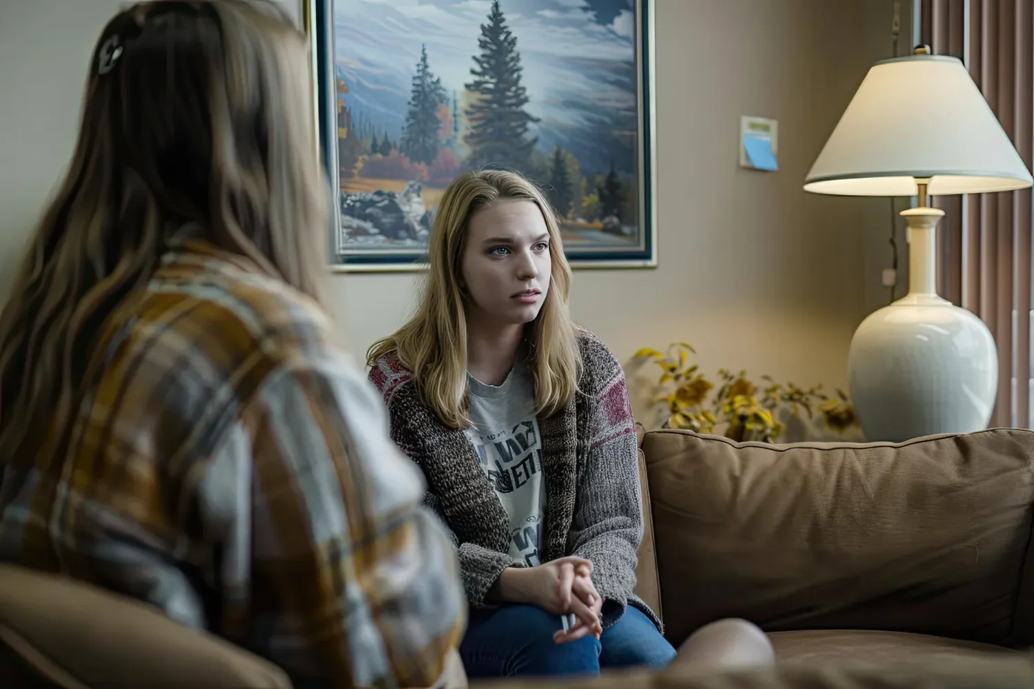 Two young women sitting on a brown couch in a living room with a painting of a mountain landscape and a white lamp in the background. One woman has blonde hair, a gray t-shirt, and a colorful cardigan. The other woman has brown hair with a clip and a plaid shirt, with her back to the camera.