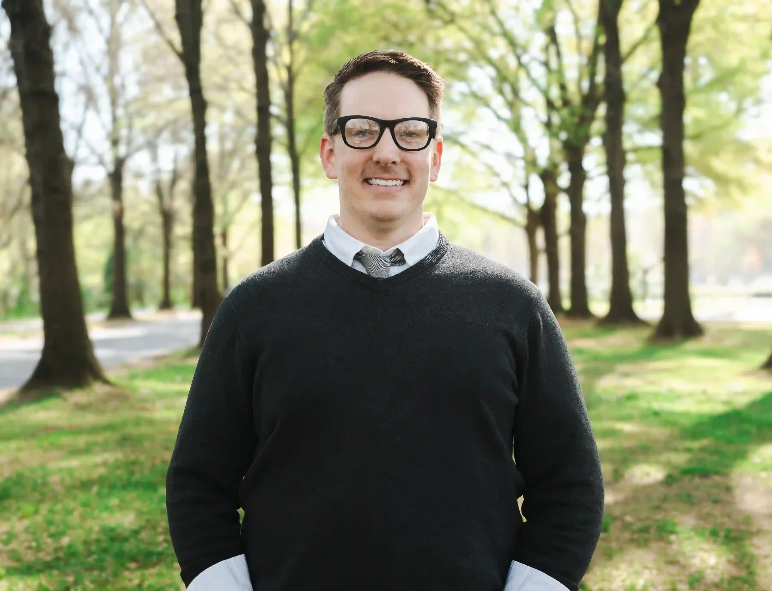 A smiling young man wearing glasses, a white shirt, gray tie, and a black sweater, standing outdoors in a park with trees and green grass.
