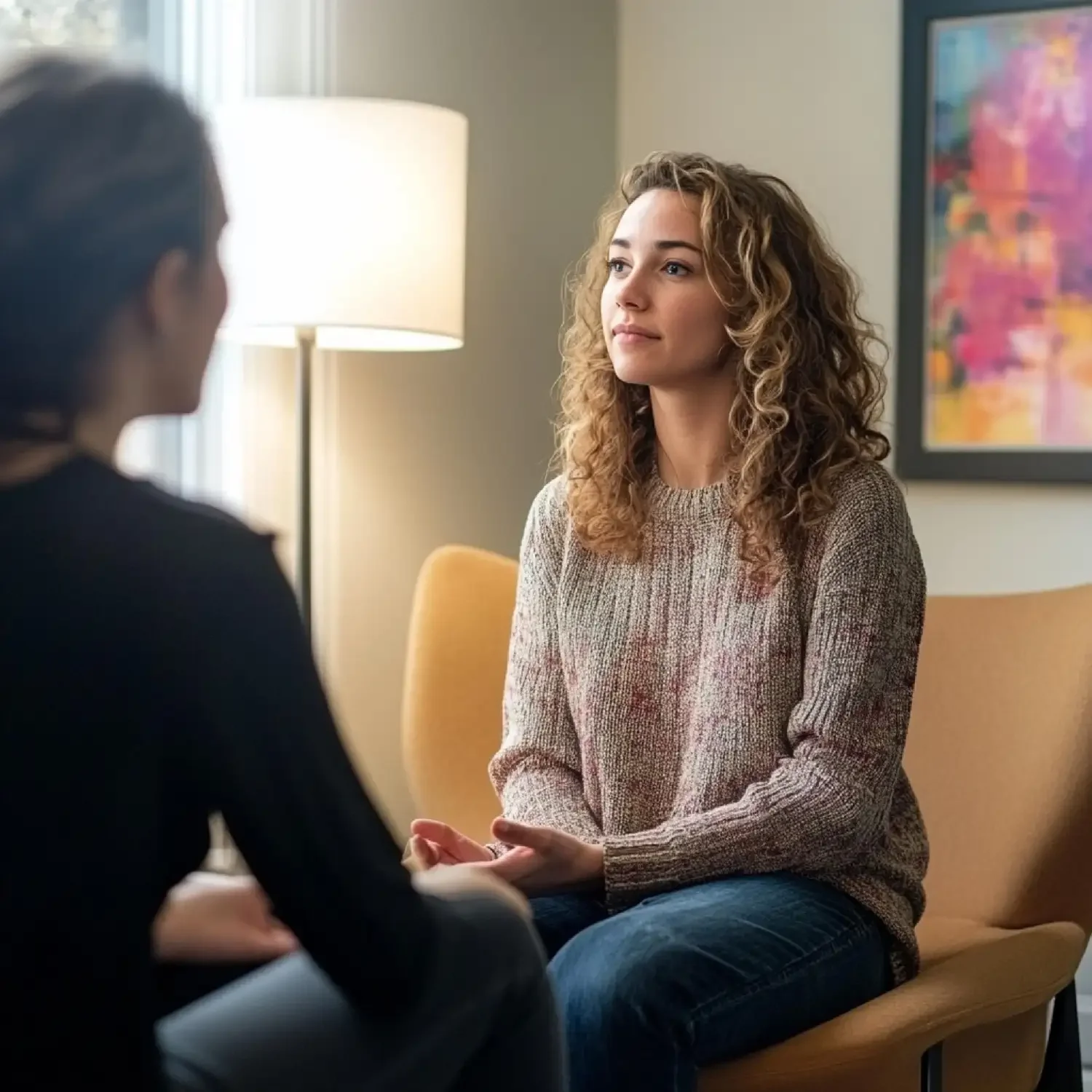 A woman with curly blonde hair sitting on a yellow chair, engaged in conversation with another person in a cozy, well-lit room.