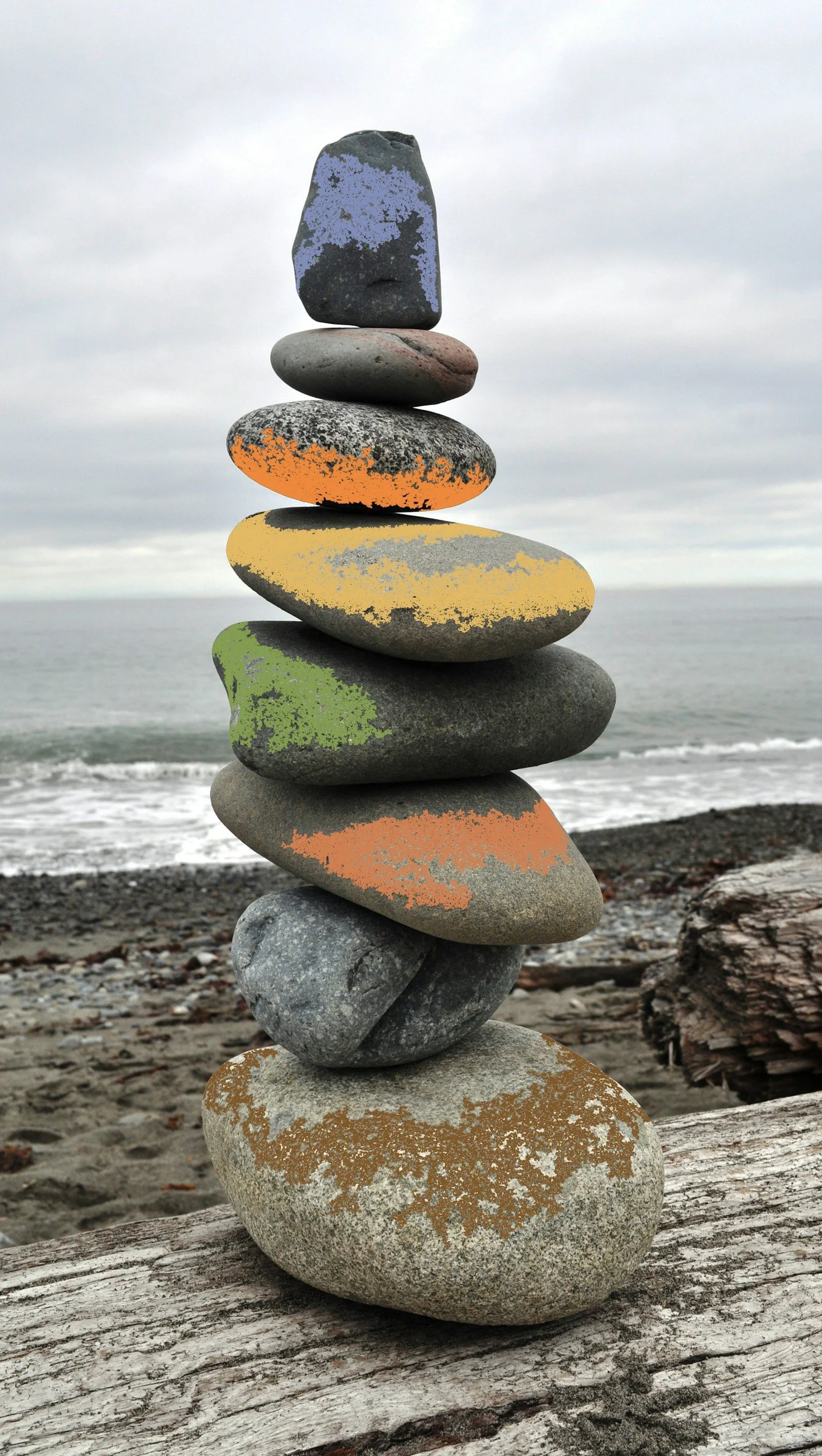 A stack of eight smooth rocks balancing on a weathered driftwood log on a rocky beach. The sky is overcast, and the ocean is visible in the background.